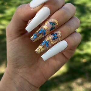 Puede incluir: Uñas acrílicas blancas largas en forma de ataúd con detalles de pan de oro y mariposas azules.