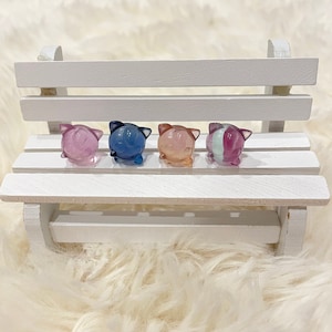 May include: Four small, colorful, resin figurines shaped like cats, sitting on a white wooden bench. The figurines are in shades of pink, blue, and purple.