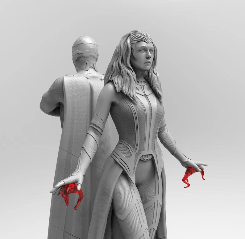 Op de afbeelding: Een grijs en wit beeldje van een vrouw met lang haar en een vloeiende cape. Ze houdt een rood voorwerp in haar rechterhand. Het beeldje staat naast een grijs en wit beeldje van een man met een cape.