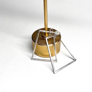 May include: A pair of silver-toned, square-shaped earrings. The earrings are displayed on a gold-colored jewelry stand against a white background. The earrings have a minimalist, geometric design.
