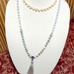 May include: A light blue and white beaded necklace with a purple bead and a grey tassel. The necklace is displayed on a white mannequin.