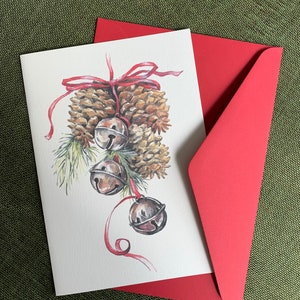 Peut inclure: Une carte de Noël illustrant des pommes de pin et des cloches attachées avec un ruban rouge. La carte est vierge à l'intérieur et est livrée avec une enveloppe rouge.