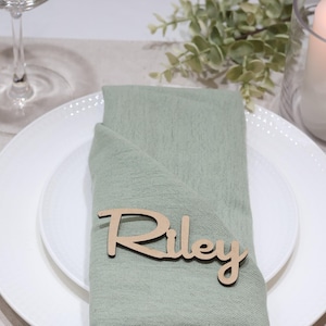 May include: A light green fabric napkin folded on a white plate, featuring a custom wooden name tag with the name Riley in a cursive script font. The place setting includes gold-toned cutlery, a glass, and a white pillar candle in a glass holder.