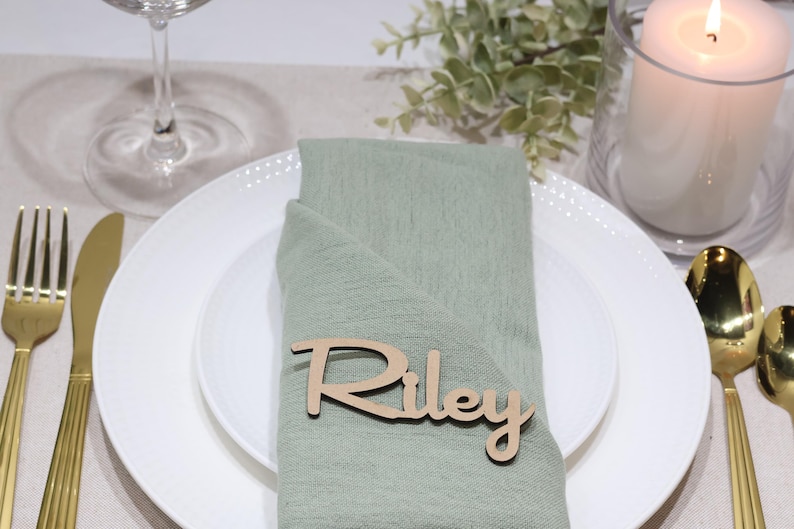 May include: A light green fabric napkin folded on a white plate, featuring a custom wooden name tag with the name Riley in a cursive script font. The place setting includes gold-toned cutlery, a glass, and a white pillar candle in a glass holder.