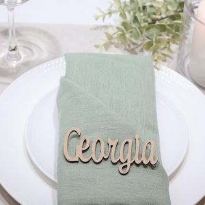 May include: A light green fabric napkin folded on a white plate, featuring a wooden name tag with the text Georgia in a cursive font. The setting includes gold-toned cutlery, a clear glass, and a white pillar candle in a glass holder.