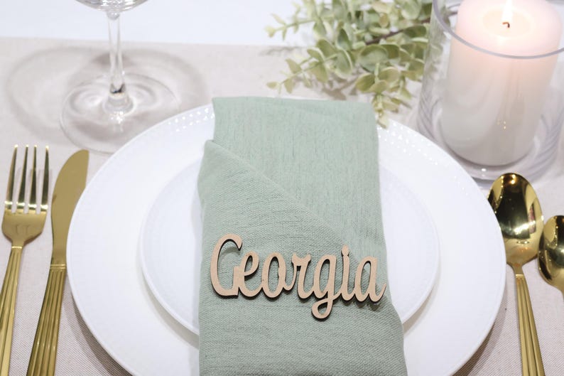 May include: A light green fabric napkin folded on a white plate, featuring a wooden name tag with the text Georgia in a cursive font. The setting includes gold-toned cutlery, a clear glass, and a white pillar candle in a glass holder.