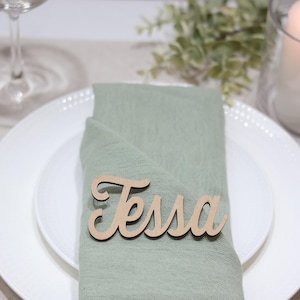 May include: A light green fabric napkin folded on a white plate, featuring a wooden name tag with the name Tessa written in a cursive script font. The place setting includes gold-toned cutlery, a glass, and a white pillar candle nearby.