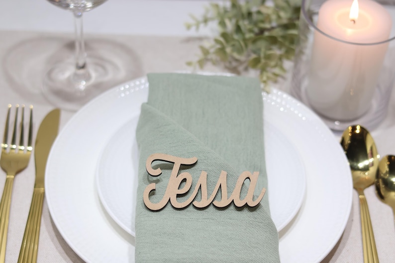 May include: A light green fabric napkin folded on a white plate, featuring a wooden name tag with the name Tessa written in a cursive script font. The place setting includes gold-toned cutlery, a glass, and a white pillar candle nearby.