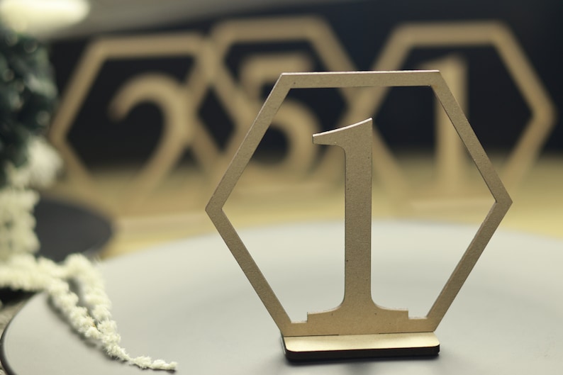 May include: A wooden table number in the shape of a hexagon with the number "1" cut out. The table number is on a wooden stand and is a light brown color.