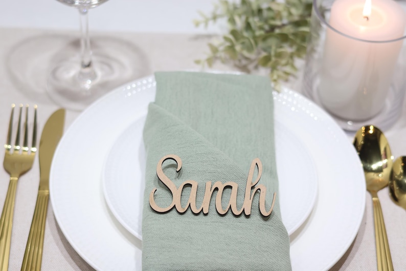 May include: A light brown wooden name tag with the name Sarah in a cursive script font, placed on a sage green fabric napkin. The napkin rests on a white ceramic plate, accompanied by gold-toned cutlery and a white pillar candle in a glass holder.