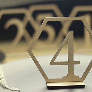 May include: A wooden table number in the shape of a hexagon with the number "4" cut out in the center. The table number is on a wooden base and is sitting on a gray surface.