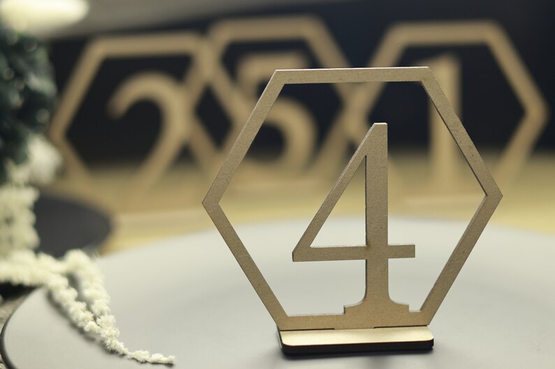 May include: A wooden table number in the shape of a hexagon with the number "4" cut out in the center. The table number is on a wooden base and is sitting on a gray surface.