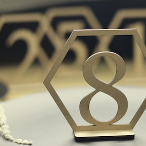 May include: Wooden table number with the number 8 cut out in the center. The number is in a hexagonal frame. The table number is on a gray surface.