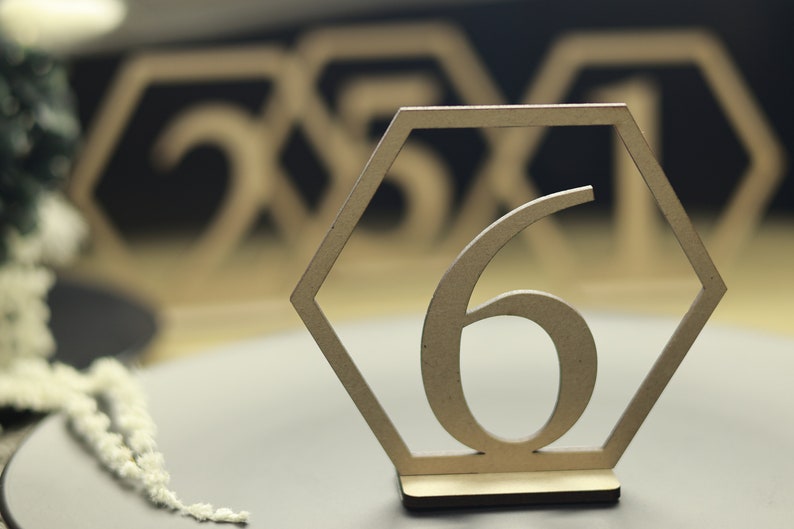 May include: A wooden table number in the shape of a hexagon with the number 6 in the center. The table number is on a gray surface with other table numbers in the background.