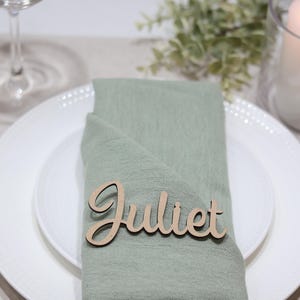 May include: A light green fabric napkin folded on a white plate, featuring a laser-cut wooden name tag with the name Juliet in a cursive script font. The setting includes gold-toned cutlery and a white pillar candle in a glass holder.