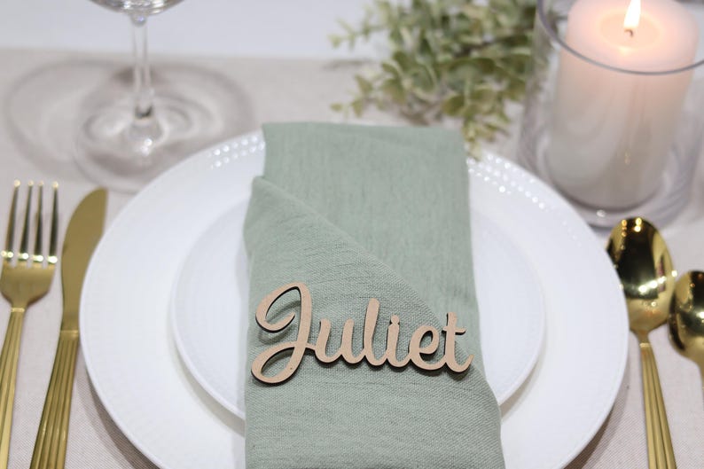 May include: A light green fabric napkin folded on a white plate, featuring a laser-cut wooden name tag with the name Juliet in a cursive script font. The setting includes gold-toned cutlery and a white pillar candle in a glass holder.