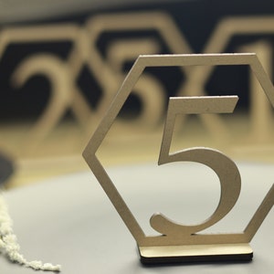 May include: Wooden table number with the number 5 cut out in the center. The number is in a hexagonal frame. The table number is on a gray surface.