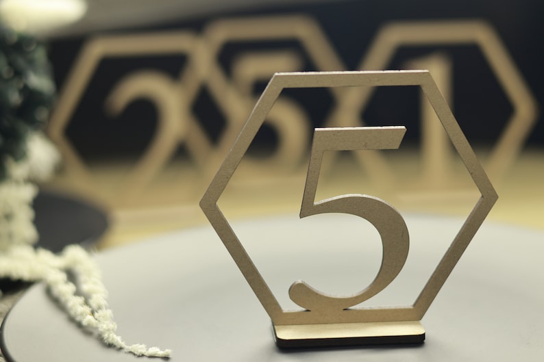 May include: Wooden table number with the number 5 cut out in the center. The number is in a hexagonal frame. The table number is on a gray surface.