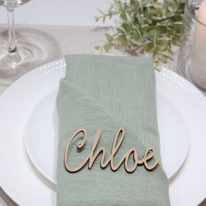 May include: A light wood laser-cut name tag featuring the name Chloe in a cursive script font, placed on a sage green fabric napkin folded over a white ceramic plate. The name tag is made of thin, flat wood with a natural light brown finish.