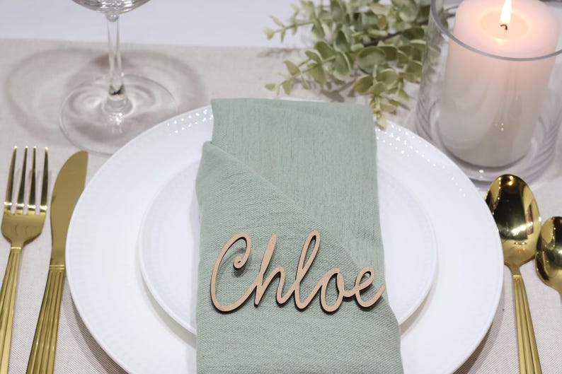 May include: A light wood laser-cut name tag featuring the name Chloe in a cursive script font, placed on a sage green fabric napkin folded over a white ceramic plate. The name tag is made of thin, flat wood with a natural light brown finish.