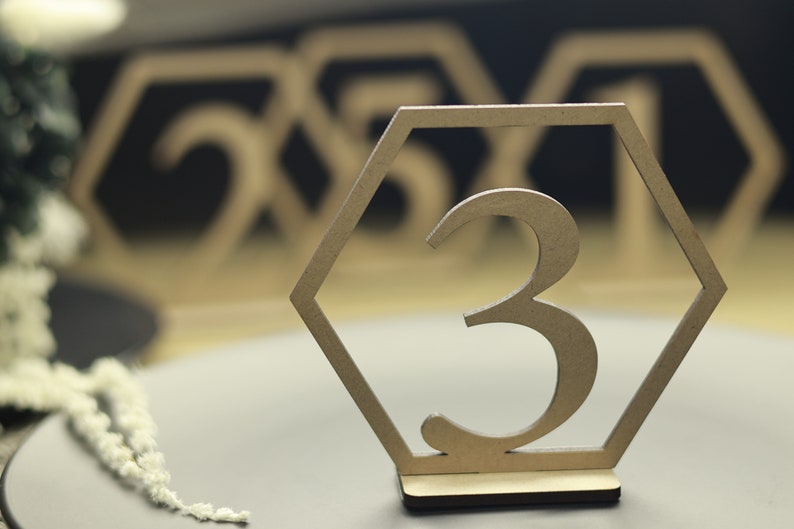 May include: A wooden table number with a hexagonal frame and the number "3" cut out in the center. The table number is on a gray surface.