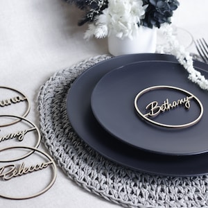 May include: Four wooden place card holders with the names "Justina", "Tommy", "Rebecca", and "Bethany" written in cursive. The place cards are on a dark blue plate with a gray crocheted placemat.