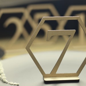 May include: A wooden table number in the shape of a hexagon with the number 7 cut out of the center. The table number is on a wooden base.
