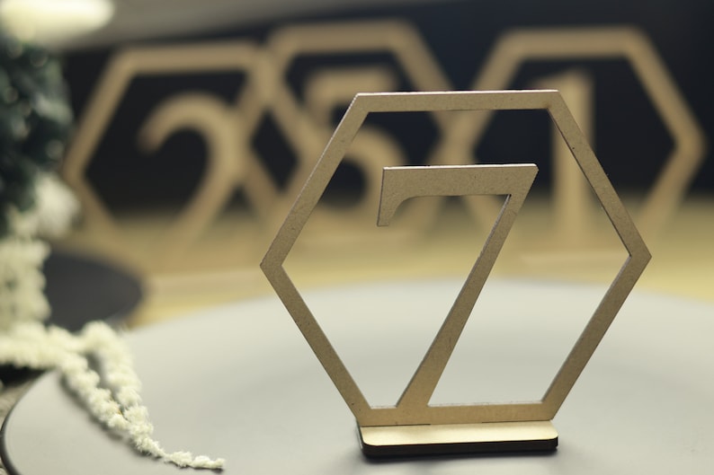 May include: A wooden table number in the shape of a hexagon with the number 7 cut out of the center. The table number is on a wooden base.