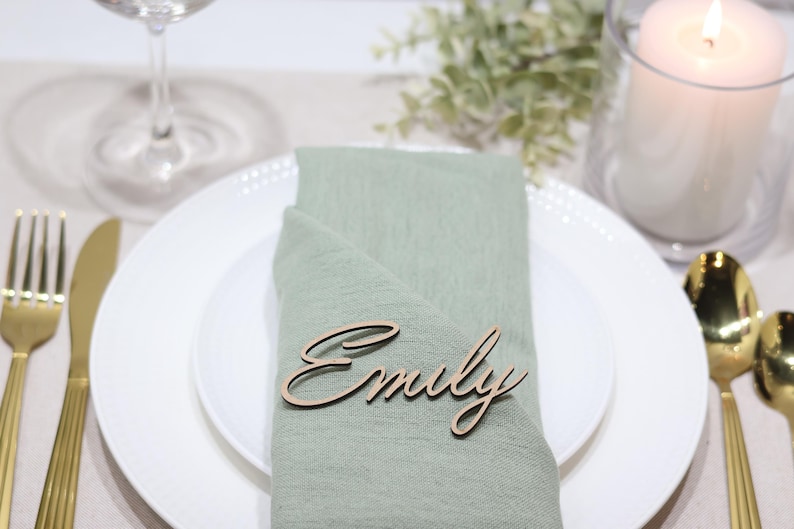May include: A laser-cut wooden name tag featuring the name Emily in a cursive script font, placed on a sage green fabric napkin folded over a white ceramic dinner plate, accompanied by gold-toned flatware and a white pillar candle.