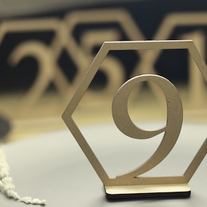 May include: A wooden table number in the shape of a hexagon with the number 9 cut out. The table number is on a gray surface.