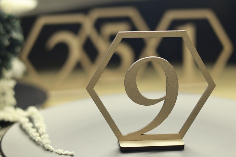 May include: A wooden table number in the shape of a hexagon with the number 9 cut out. The table number is on a gray surface.