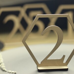 May include: A wooden table number in the shape of a hexagon with the number "2" cut out. The table number is on a gray surface.
