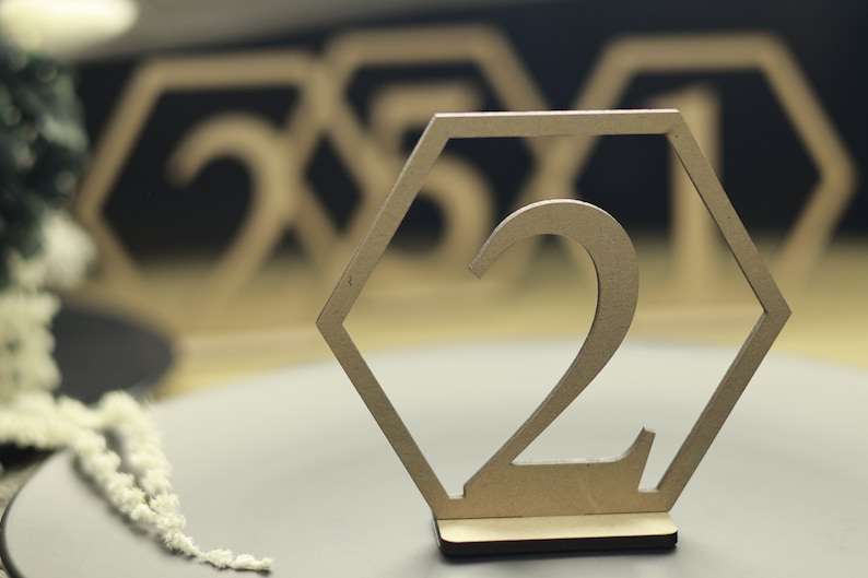 May include: A wooden table number in the shape of a hexagon with the number "2" cut out. The table number is on a gray surface.