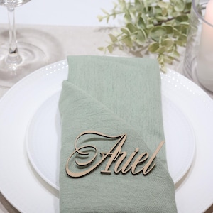 May include: A light sage green fabric napkin folded on a white ceramic plate, featuring a laser-cut wooden name tag with the name Ariel in a cursive script font. The setting includes gold-toned cutlery, a glass, and a white pillar candle.