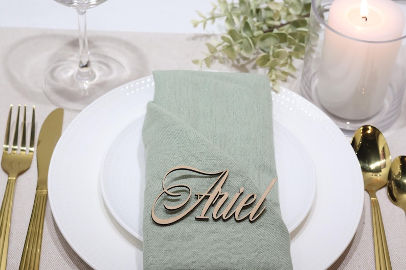 May include: A light sage green fabric napkin folded on a white ceramic plate, featuring a laser-cut wooden name tag with the name Ariel in a cursive script font. The setting includes gold-toned cutlery, a glass, and a white pillar candle.