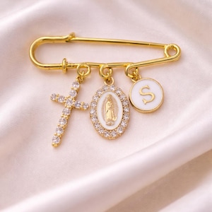 May include: Gold-coloured safety pin with three charms: a crystal-studded cross, an oval pendant with a religious figure, and a white disc with a gold letter "S". The pin is displayed on white satin fabric.