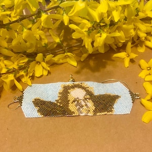 May include: A beaded bracelet featuring a lion design on a light blue background. The lion is rendered in brown, tan, and cream beads. The bracelet has gold-coloured chain and clasp elements. Yellow flowers are visible in the background.