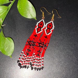 Peut inclure: Une paire de longues boucles d'oreilles pendantes composées de petites perles de rocaille dans une palette de couleurs rouge, noir et blanc. Elles présentent une section supérieure en losange géométrique avec un motif, prolongée par des franges.