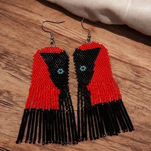 Peut inclure: Une paire de boucles d'oreilles en perles au design saisissant. Les boucles d'oreilles présentent un motif rouge et noir avec une frange de perles noires. Chaque boucle d'oreille a un petit accent de perle turquoise. Les boucles d'oreilles sont présentées sur une surface en bois.