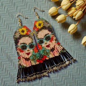 Puede incluir: Un par de pendientes de cuentas con el retrato de una mujer con gafas de sol y un tocado floral. Los pendientes tienen flecos negros y ganchos dorados. El diseño incorpora cuentas rojas, amarillas, azules y negras.