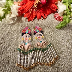 Può includere: Un paio di orecchini pendenti in perline raffiguranti il ritratto di una persona con capelli scuri, un copricapo floreale e sopracciglia marcate. Gli orecchini sono realizzati con perline di vetro multicolori, con frange verdi, oro e trasparenti.