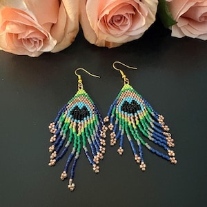 Peut inclure: Une paire de boucles d'oreilles en perles avec un motif de plume de paon. Les boucles d'oreilles présentent un dessus triangulaire avec un motif noir, bleu et vert, et de longues mèches perlées bleues, vertes et grises. Des crochets et des accents dorés complètent le design.