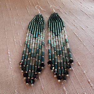 Peut inclure: Une paire de longues boucles d'oreilles pendantes composées de perles de verre cylindriques en dégradé. Les perles passent d'un vert forêt profond en haut à une teinte transparente au milieu, terminées par des perles noires à facettes.