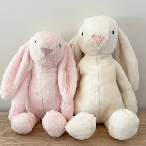 May include: Two plush bunny toys, one pink and one white, sitting side by side on a wooden surface.