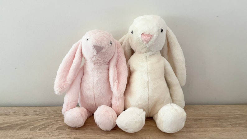 May include: Two plush bunny toys, one pink and one white, sitting side by side on a wooden surface.