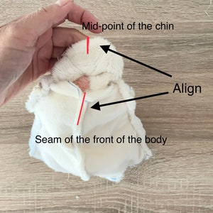 May include: A white plush fabric piece with a seam down the front. The text "Seam of the front of the body" is below the seam. The text "Mid-point of the chin" is above the fabric piece with a red line pointing to the top of the fabric piece. The text "Align" is above the fabric piece with a black line pointing to the seam.