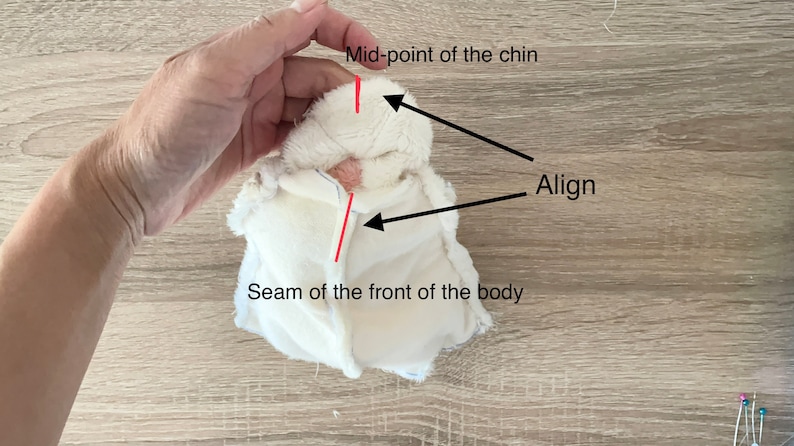 May include: A white plush fabric piece with a seam down the front. The text "Seam of the front of the body" is below the seam. The text "Mid-point of the chin" is above the fabric piece with a red line pointing to the top of the fabric piece. The text "Align" is above the fabric piece with a black line pointing to the seam.