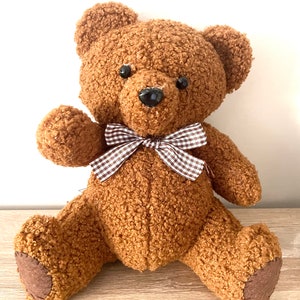 May include: A brown teddy bear with a gingham bow tie. The bear is sitting on a light brown surface.