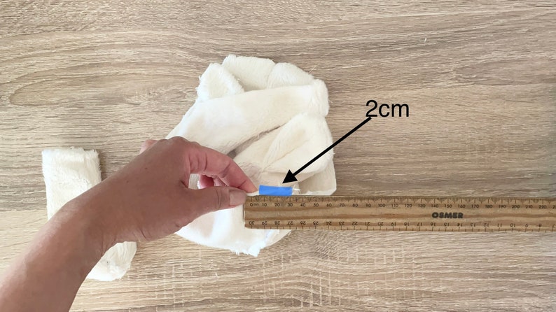 May include: White fabric with a blue tape marker measuring 2 cm in length. A wooden ruler is used to measure the fabric.