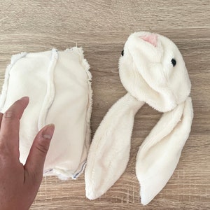 May include: A white plush bunny head with long ears and black eyes. A white fabric rectangle with a seam down the middle is also shown.